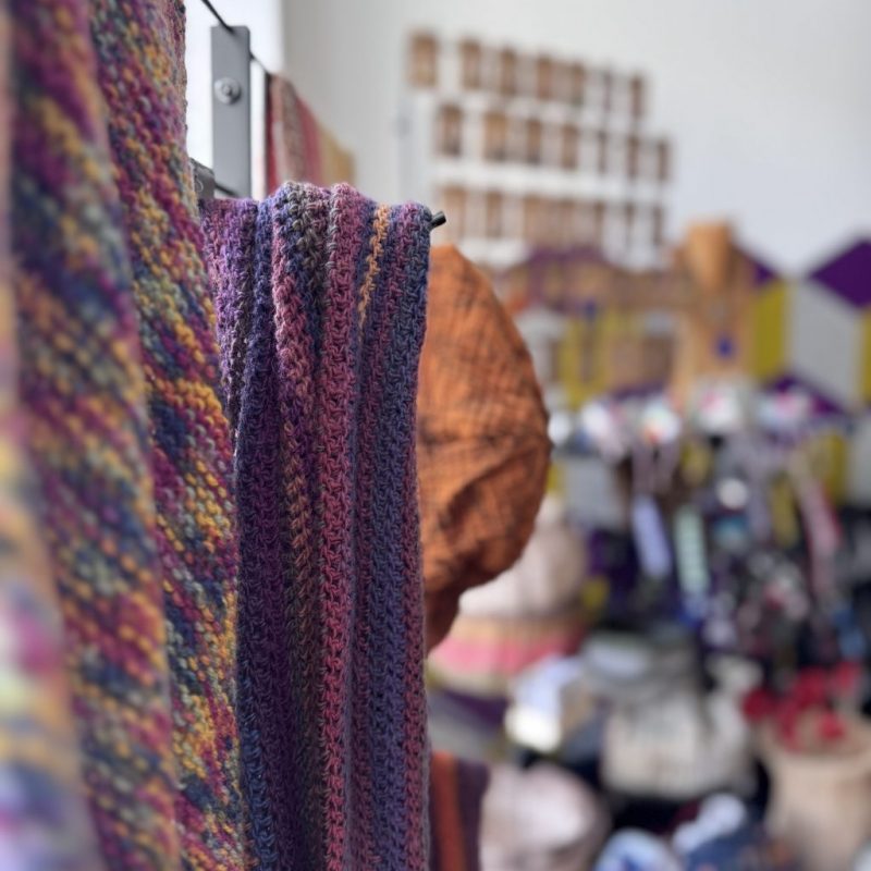 Im Vordergrund gestrickte Schals, Stoffmützen, im Hintergrund verschwommen ein Verkaufsstand mit Schmuck, Taschen, Deko
