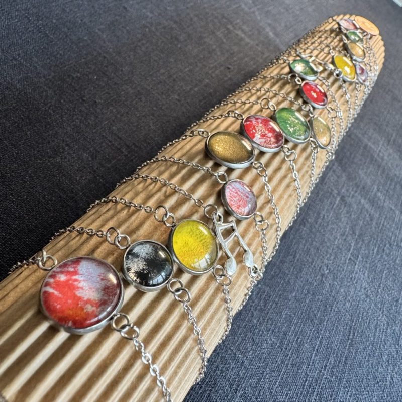 mehrere Armbänder mit silberfarbenen Ketten und Lünetten gefüllt mit buntem Papier hinter Glascabochons, angeordnet auf einer Rolle aus Wellpappe, der Hintergrund ist dunkelgrau