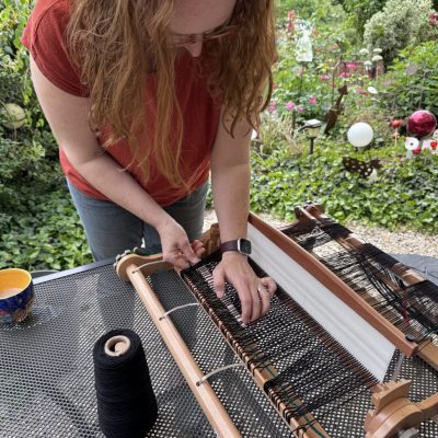 Webrahmen aufsetzen 2025 @StitchWeaveSmile Frau mit langen Haaren steht über einen Webrahmen gebeugt im Garten und befestigt schwarze Fäden.