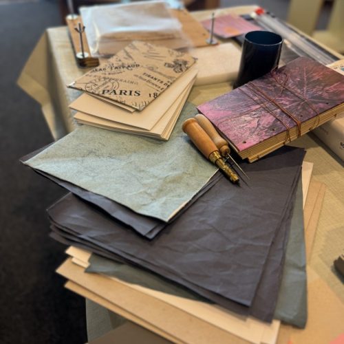 Buchbinden @StitchWeaveSmile Material für Buchinden auf einem Tisch. Es sind dunkelblaues Papier, ein mit Stoff bezogener Buchdeckel sowie ein noch zu bindendes Buch mit lila Cover zu sehen, daneben eine Ahle und ein Papierbohrer.