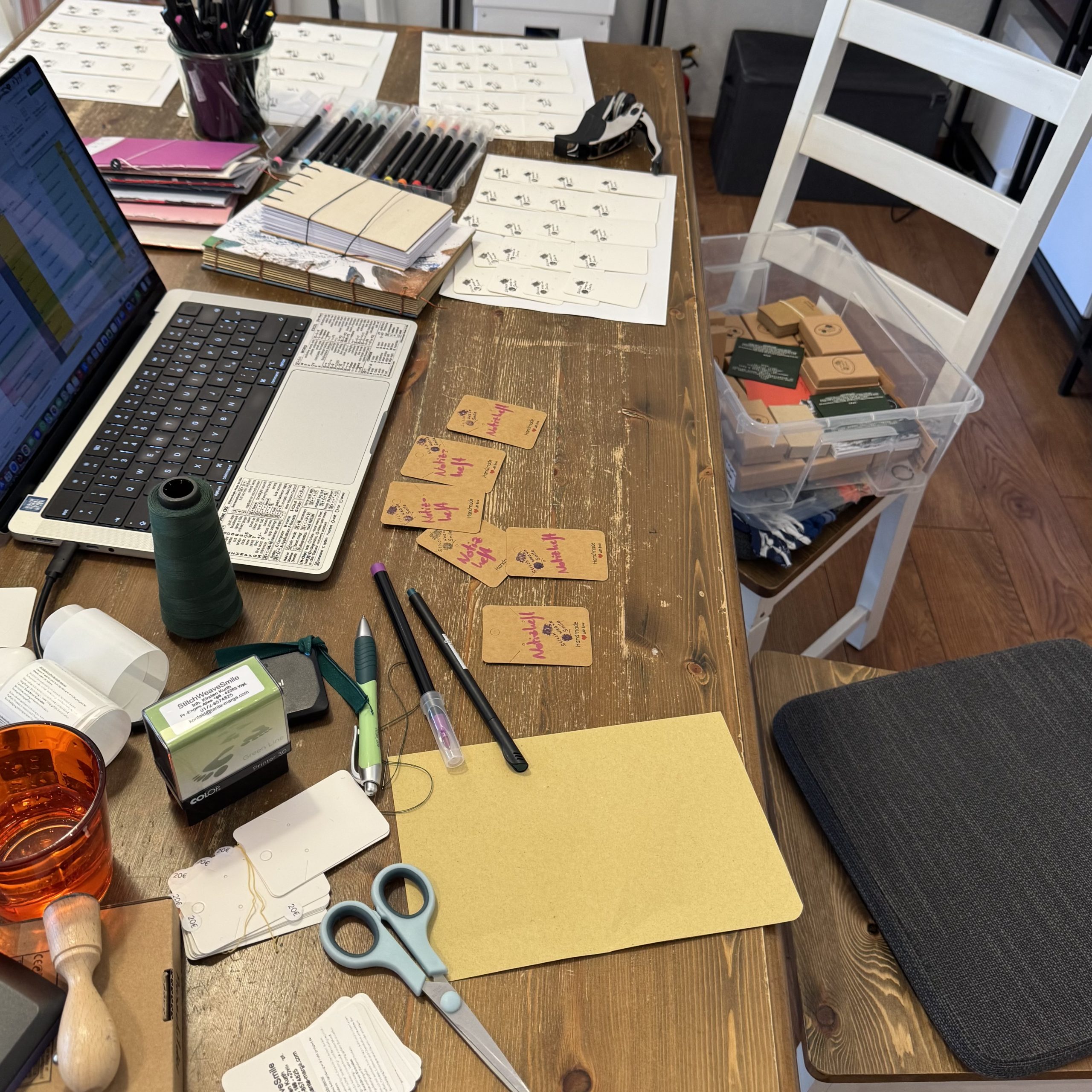 Auf einem dunklen Holztisch steht ein Laptop.Daneben liegen gestempelte weiße Schmuckkarten, handbeschriebene Preisschilder sowie allerlei Utensilien.